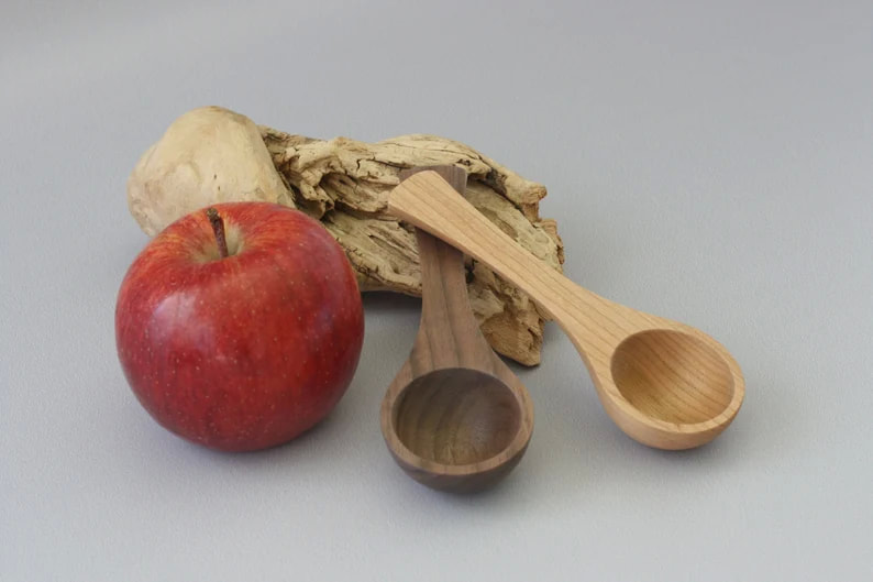 Walnut and cherry wood coffee scoops for organic coffee on a gray background, with a driftwood prop and and apple for scale.