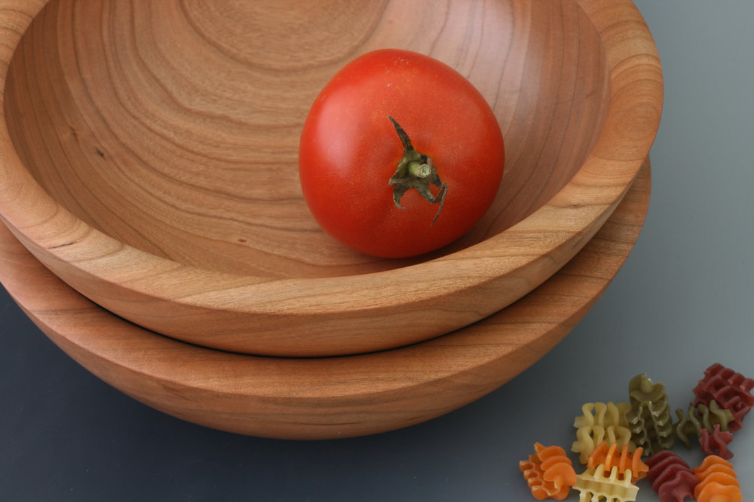 Two cherry wood bowls on a gray background with a tomato and tricolor pasta for scale.