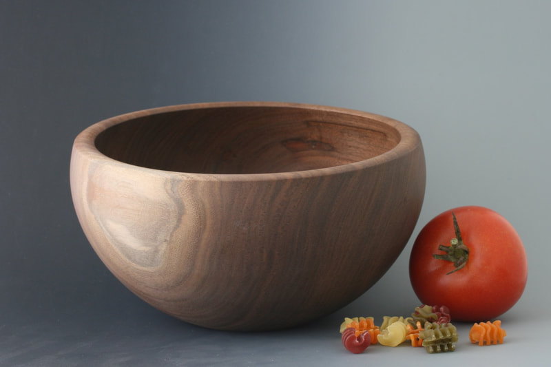 Black walnut wood bowl against a gray gradient backdrop with tricolor pasta and a tomato for scale.