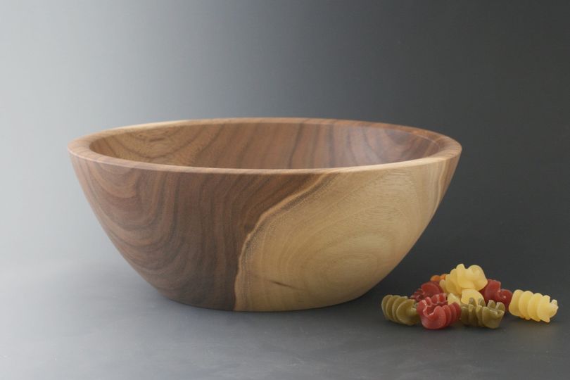 Black walnut wood bowl against a gray gradient backdrop with tricolor pasta and a tomato for scale.
