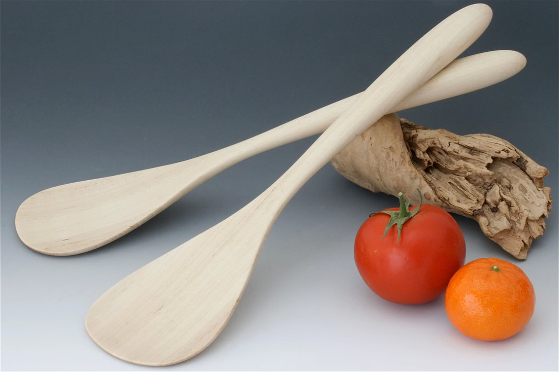 Maple wood salad serving paddles resting on a driftwood prop on a gray gradient background and with a tomato and clementine fruit for scale.