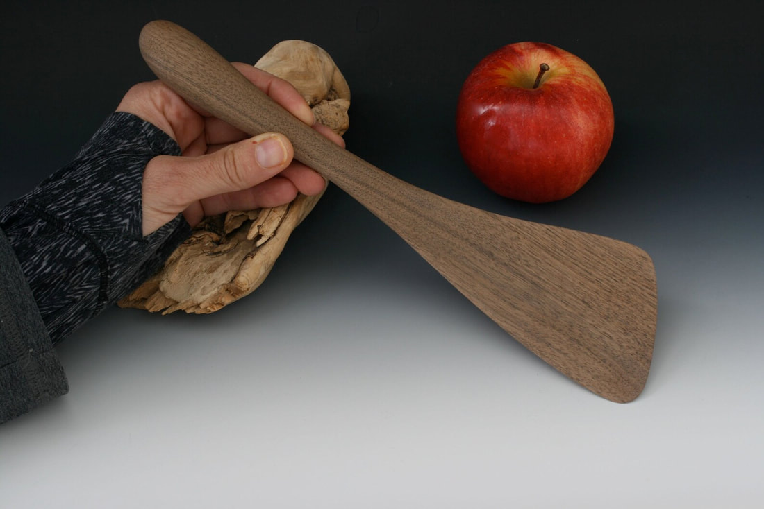 PictureSmall walnut wood spatula photographed on a gray gradient background with an apple and being held in hand for scale.