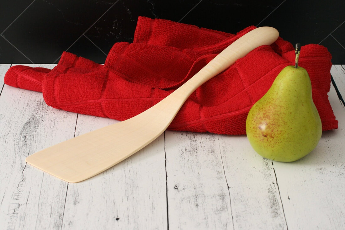 Maple wood spatula flipper turner photographed on a rustic whitewashed countertop with a black marble tile backsplash and with a pear and red dish towel for scale.