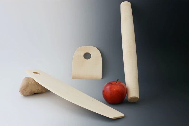Maple wood counter scraper, rocker-style pizza cutter, and rolling pin in maple wood on a gray gradient background and apple for scale.