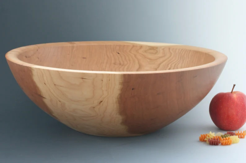 Deep wooden salad bowl on a gray gradient background with an apple and dry radiatori pasta for scale.