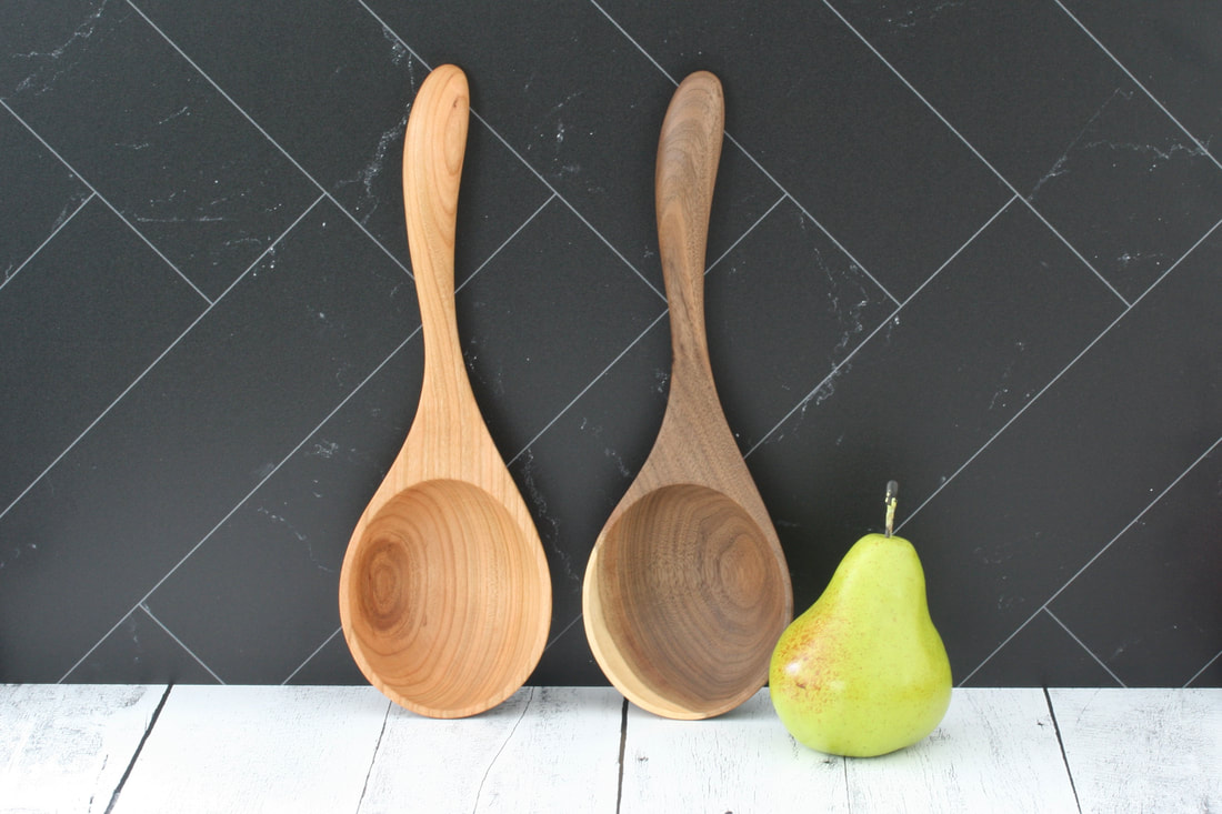 Cherry wood ladle and black walnut wood ladle on whitewashed countertop and black marble back splash with pear for scale.