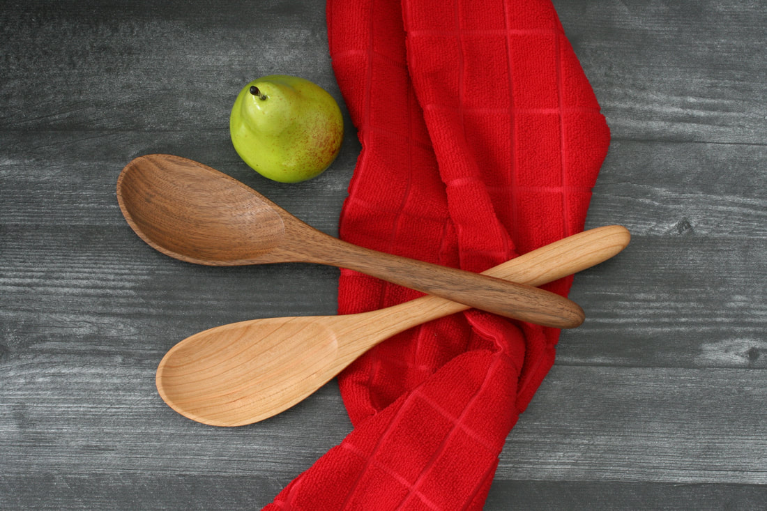 Wooden spoons with angled bowl in black walnut and black cherry against a dark wood countertop, red hand towel and a pear used for scale.