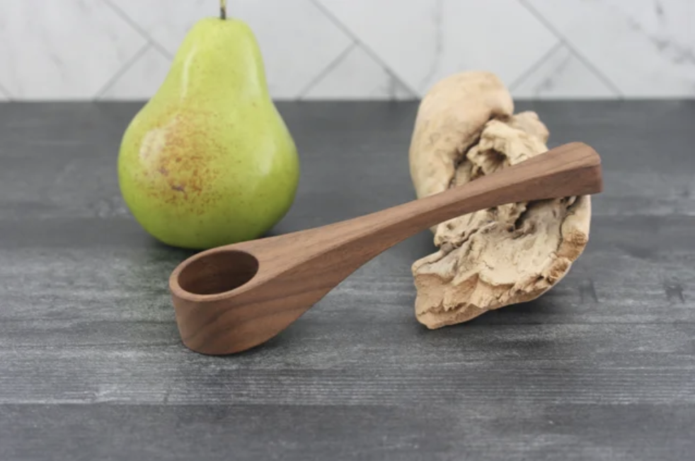 Angled shot of a black walnut pipe style coffee scoop on a distressed gray tabletop and marble backsplash with a greeen pear for scale.