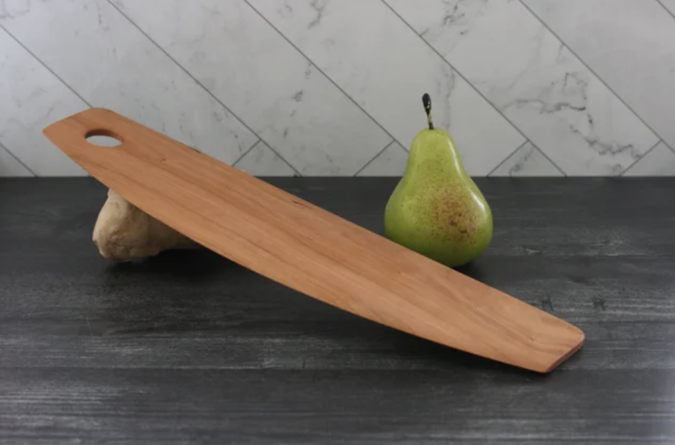 Side angled photograph of a Cherry Wood Rocker Style Pizza Cutter on a gray countertop and marble backsplash with a pear for scale.