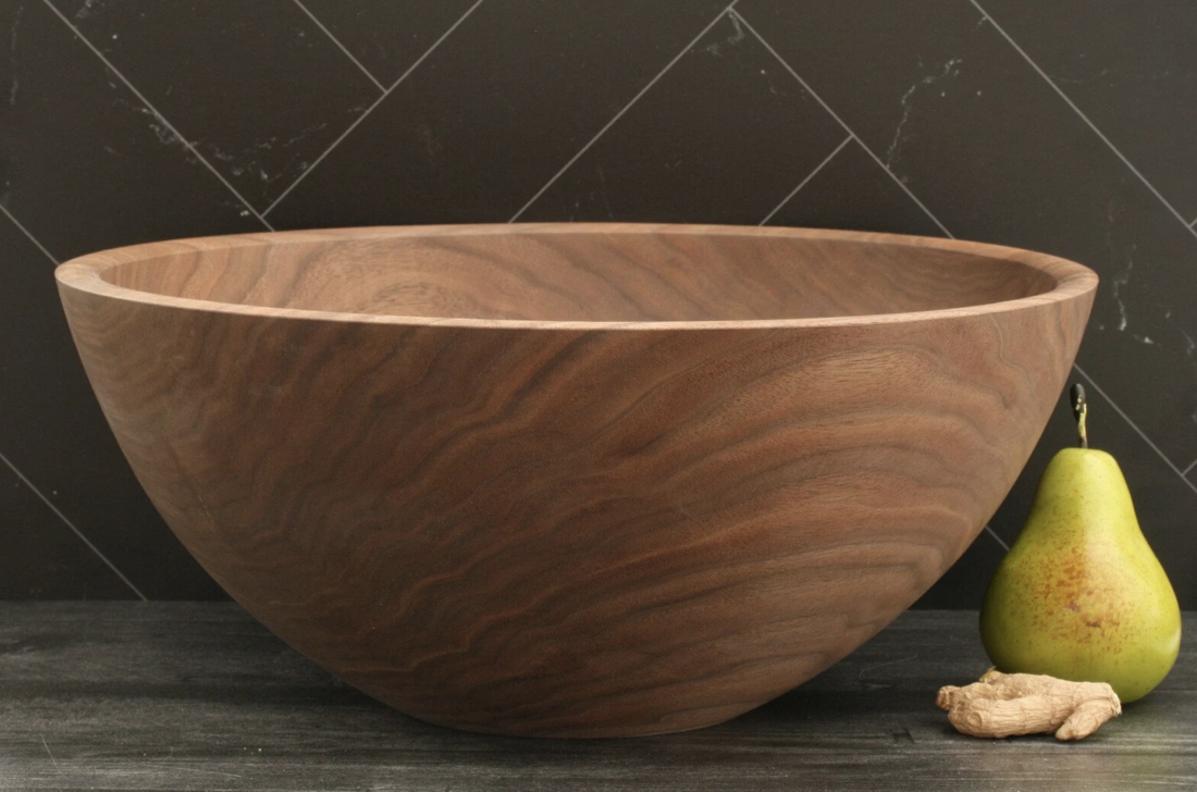 Large handmade walnut wood bowl on a dark barnwood counter top and a black tile back splash and pear for scale.